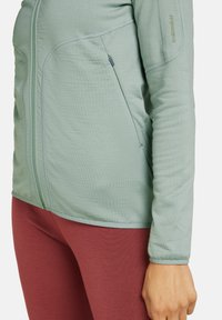 Veste longue à manches longues vert clair avec une finition texturée et une poche zippée. Associée à des leggings ajustés bordeaux.