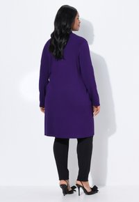 Frau mit langen schwarzen Haaren, die einen knielangen lila Mantel, schwarze Hosen und schwarze High Heels mit Schleifen trägt, steht vor einer weißen Wand.