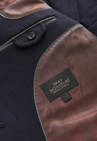 Abrigo de lana azul marino con acentos grises, que cuenta con un bolsillo y un forro liso de color burdeos con patrón, etiquetado como "NEXT SIGNATURE PREMIUM TAILORING."