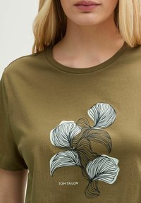 T-shirt en coton vert olive avec un col rond présentant un design floral abstrait noir et blanc. "TOM TAILOR" imprimé en blanc en dessous.
