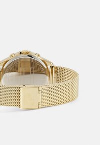 Montre avec un bracelet en maille dorée, un boîtier rond et trois boutons, présentant un dos en verre affichant un cadran minimaliste et un joli gravage de la marque.