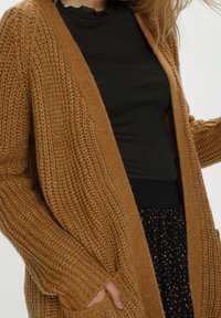 Sweter rozpinany na drutach w ciepłym brązowym kolorze z otwartym przodem, ściągaczami na mankietach i dwoma przednimi kieszeniami, noszony na czarnej, teksturowanej bluzce.