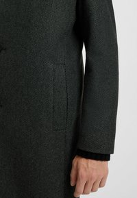 Mørkegrøn herringbone-frakke med struktureret stof, strømlinet form, klaplomme og ribbet sort manschetdetalje.