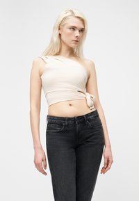 Ξανθιά γυναίκα που φοράει ένα μπεζ crop top με έναν ώμο και μαύρο τζιν με ψηλή μέση, στέκεται μπροστά σε απλό λευκό φόντο.