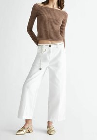 Henkilö, jolla on päällä ruskea ribattu pois olkapäältä oleva crop top, valkoiset leveät housut, joissa on ketjuhihna, ja kultaiset kärjetön slingback-kengät.