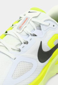 Λευκό αθλητικό παπούτσι με γκρι και νέον κίτρινες λεπτομέρειες, με υφασμένο επάνω μέρος, διακριτικό λογότυπο Nike και επενδεδυμένο κολάρο για υποστήριξη.