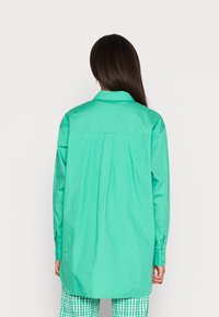 Camisa de mangas longas em verde brilhante, feita de tecido suave. Apresenta um colarinho tradicional e um pregueado nas costas. Combinada com calças de padrão quadriculado.