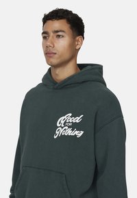 Felpa verde in cotone con tasca frontale, caratterizzata da una stampa di testo bianca con la scritta "Good for Nothing" in un carattere stilizzato. Vestibilità classica con cappuccio regolabile.