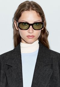 Occhiali da sole con montatura marrone scuro e lenti verdi, caratterizzati da una forma rettangolare ampia. Indossati con un dolcevita bianco e un blazer grigio.