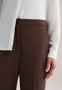 Bruine op maat gemaakte broek met een gladde textuur, voorzien van een enkele zwarte knoopsluiting, gecombineerd met een witte, geplooide blouse.