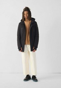 Jonge man met dreadlocks, gekleed in een zwarte hooded jack, beigemip shirt, witte broek en zwarte schoenen, staande tegen een effen grijze achtergrond.