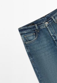 Jeans in denim blu piegati che mostrano la tasca anteriore, la vita, i passanti per la cintura e la chiusura con bottone su uno sfondo bianco semplice.