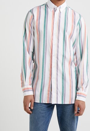 Mężczyzna noszący koszulę z długim rękawem z pionowymi paskami w kolorach zielonym, różowym, białym i czarnym, w połączeniu z niebieskimi dżinsami.