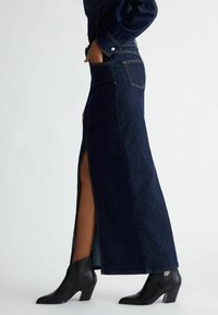 Gonna maxi in denim scuro con spacco laterale, cuciture gialle e tasche posteriori. Indossata con stivaletti neri alla caviglia, caratterizzati da punta e tacco appuntiti.