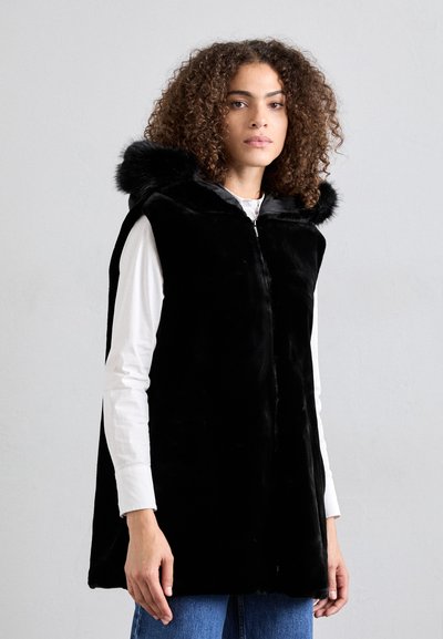 Black fur vest with a hood, featuring a smooth texture and zip closure. Worn over a white shirt, has front pockets and a relaxed fit.