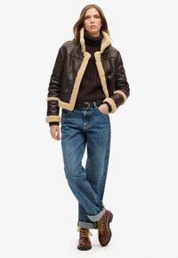 Brązowa skórzana kurtka z owczej wełny z kremowym podbiciem, ciemny prążkowany sweter, jasnoniebieskie dżinsy z podwiniętymi nogawkami oraz brązowe buty za kostkę z żółtymi sznurówkami.