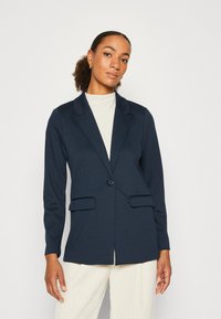 Blazer bleu marine avec revers, fermeture à un bouton, deux poches avant, coupe ajustée, fabriqué en tissu texturé. Porté par-dessus un haut clair.