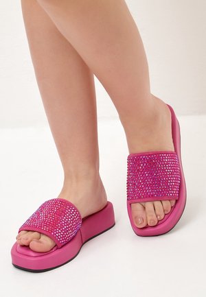 Feet wearing bright pink platform slide sandals with sparkling rhinestone-covered straps on a white background.
