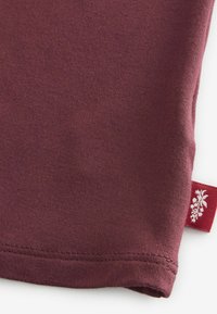 Esquina de camiseta de algodón burdeos con textura fina, dobladillo cosido y etiqueta roja con diseño floral blanco.