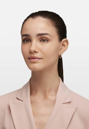 Boucles d'oreilles en or avec une surface texturée, portées avec un blazer rose clair. Peau lisse, cheveux noirs droits et maquillage naturel sont visibles.