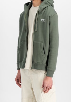 Mand iført en olivengrøn hættetrøje med lynlås foran og lommer samt Alpha Industries-logo, kombineret med beige cargobukser og en beige skjorte.