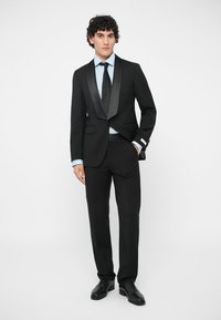 Černý smoking s saténovými klopami, spárovaný se světle modrou košilí a černou kravatou. Přiléhavý design, ušité kalhoty a lesklé černé společenské boty.