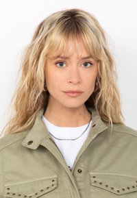 Giacca verde chiaro con borchie sulle tasche del petto, indossata su una camicia bianca. La modella ha capelli biondi ondulati di lunghezza spalle e un trucco neutro.
