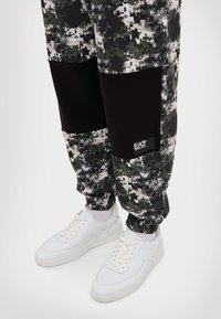 Camo-patterned black and green jogger pants with elastic cuffs and a branded label near the pocket, paired with white lace-up sneakers.