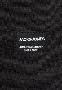 Svart tygmärke med vit text som säger "JACK & JONES" och "KVALITETSÅTTA SEDAN 1990." Rektangulär form med sydda kanter.