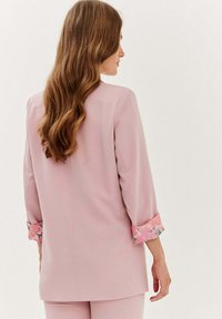 Světle růžový blazér s třičtvrtinovými rukávy, zdobený květinovým vzorem na manžetách. Hladký materiál, přiléhavý střih a minimalistický design.