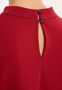 Rotes Stoffoberteil mit hohem Kragen und einer kleinen Rückenschlitzöffnung, die durch zwei Knöpfe gesichert ist. Es hat eine glatte Textur und saubere Nähte.
