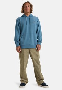 Blue cotton hoodie with a front pocket, adjustable drawstring, and embroidered logo. Paired with tan corduroy pants and blue sneakers.