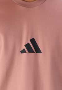 Rozā kokvilnas T-krekls ar melnu Adidas logotipu. Apaļa kakla izgriezums un gluda tekstūra izceļ tā vienkāršo dizainu.