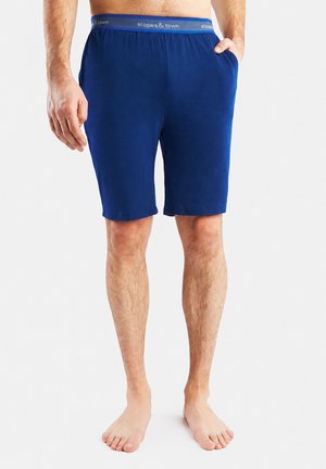 Uomo che indossa pantaloncini da casa blu fino al ginocchio con cintura "slopes & town", in piedi a piedi nudi su sfondo bianco.