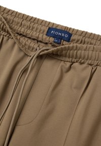 Braune Hose mit elastischem Bund und Kordelzug, die eine glatte Textur und subtile Falten aufweist, innen mit "PIOMBO" etikettiert.