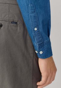 Detail na ruke osoby v modrej košeli s gombíkmi, ktorá odpočíva vedľa sivých nohavíc s malým logom Hackett blízko vrecka.