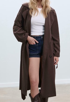Blonde Person trägt einen langen braunen Mantel über einem weißen bauchfreien Top und dunklen Jeansshorts, mit der linken Hand in der Tasche.