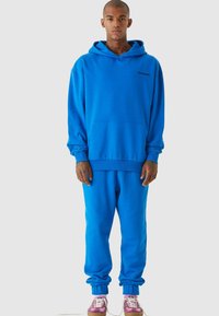 Felpa blu con tasca a marsupio e polsini a costine, abbinata a pantaloni della tuta blu coordinati. Indossato con sneaker rosa e bianche.