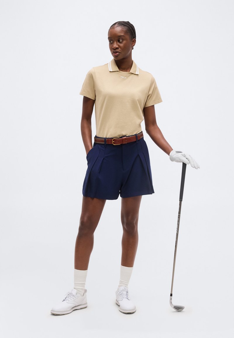 Femme en polo beige, short bleu marine, gant de golf blanc tenant un club de golf, posant devant un fond clair uni.