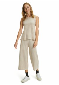 Beige ärmelloses Oberteil mit geschichtetem Saum, kombiniert mit beigen weitem Hosen, die eine weiche Textur haben, und weißen Sneakers.