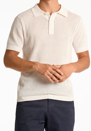 Stickad kortärmad polo, krämfärgad, med öppet vävmonteringsmönster, nedfällbar krage och ribbade ärmslut. Bärs över mörkblå byxor.