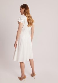 Biała sukienka do kolan z krótkimi rękawami, zwężana w talii i z plisowanym dołem. Gładki materiał z subtelnym połyskiem; noszona z beżowymi szpilkami.