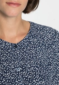T-shirt azul marinho com estampado floral branco, gola redonda e detalhe de um botão. Tecido suave com uma textura sutil, ajustado na gola.