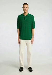 Zielona koszulka polo z krótkim rękawem i teksturowanym wzorem, zestawiona z beżowymi spodniami i czarnym obuwiem, prezentowana na stojącym modelu.