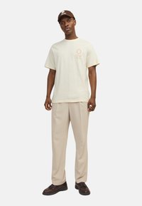 Lys beige kortærmet t-shirt med trykt logo; parret med brune bukser og brune sko. En brun kasket fuldender outfittet.