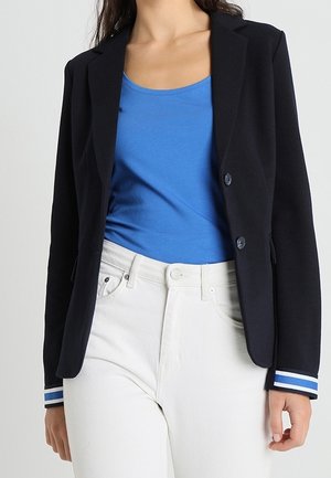 Kobieta nosząca czarną marynarkę z niebiesko-białymi paskowanymi mankietami, niebieską koszulkę na ramiączkach i białe, wysokopodcięte dżinsy.