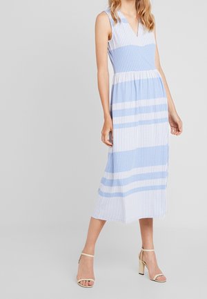 Vrouw draagt een mouwloze blauw-wit gestreepte midi-jurk, gecombineerd met crèmekleurige hakken, staand tegen een neutrale achtergrond.