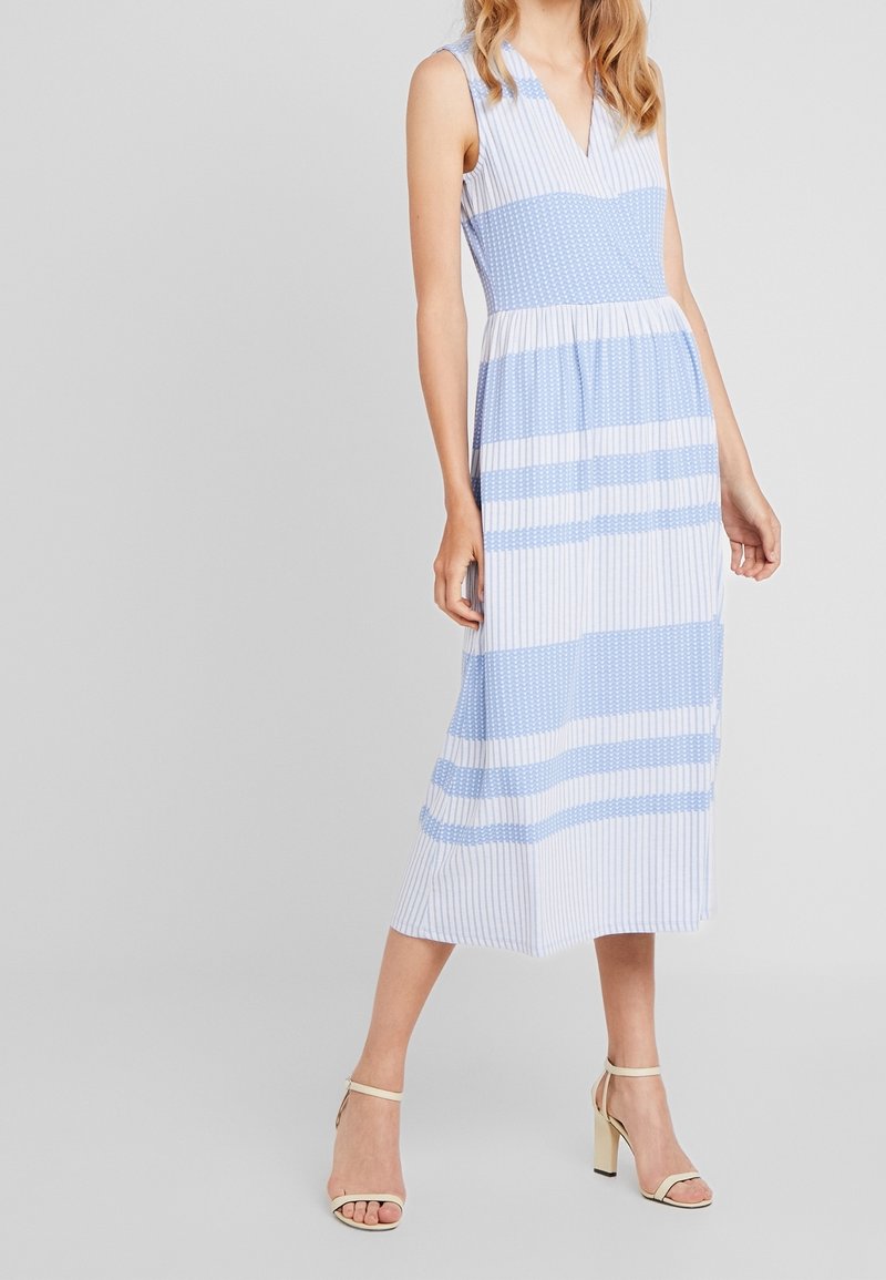 Femme portant une robe midi sans manches rayée bleu et blanc, associée à des sandales à talons hauts couleur crème, debout devant un fond neutre.
