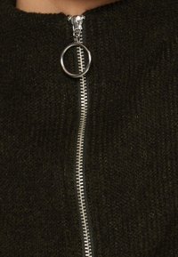 Dunkelgrüner Strickpullover mit hohem Kragen, ausgestattet mit einem metallischen Reißverschluss, einem runden Zippergriff und einem strukturierten Stoffmuster.
