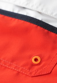 Tissu rouge à texture lisse, avec une bordure bleu marine et un œillet décoratif orange portant l'inscription "SUPERDRY" en relief. Couture détaillée visible.